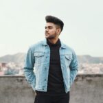 Stylish young male model in a denim jacket with a cityscape backdrop, showcasing modern fashion.